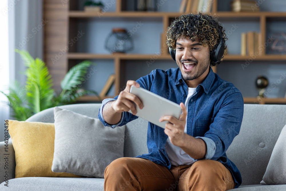 A young man with curly hair sits on a couch, engrossed in playing a game on his tablet. He wears headphones and appears excited.