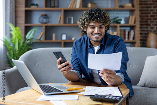 A happy man examines a bill while using his smartphone and laptop, working on his finances from home.