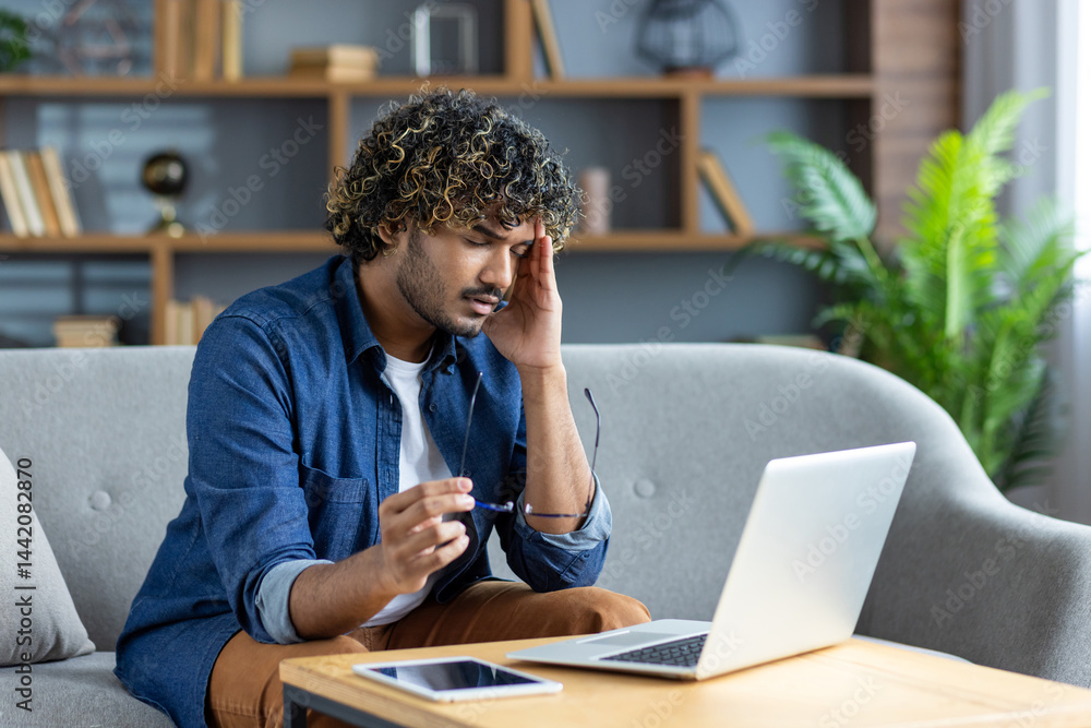 A young man, possibly experiencing a headache, sits near a laptop computer in a living room environment.