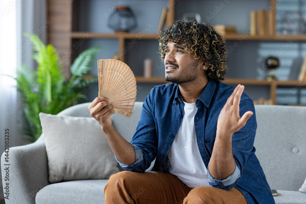 A young man with curly hair sits on a couch, holding a fan, looking uncomfortably warm. He is expressing feeling hot.