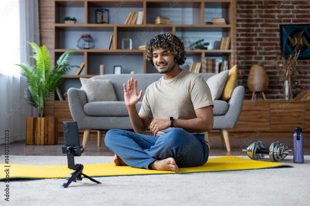 A smiling man waves while video conferencing at home during his fitness routine. The scene includes a yoga mat and dumbbells.