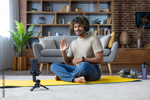 Obraz na płótnie A smiling man waves while video conferencing at home during his fitness routine. The scene includes a yoga mat and dumbbells. w salonie