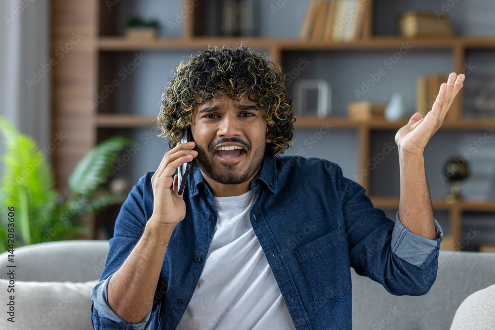 A man in a blue shirt, on a couch, is talking on his phone while gesturing with his hand, looking annoyed.