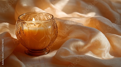A lit candle in an amber glass holder sits on a beige fabric.  Warm, soft light illuminates the scene.
