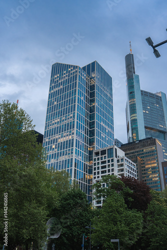 Obraz na płótnie Modern skyscrapers, including the Commerzbank Tower, rise above lush green trees in Frankfurt, Germany, under an overcast sky during dusk. w salonie