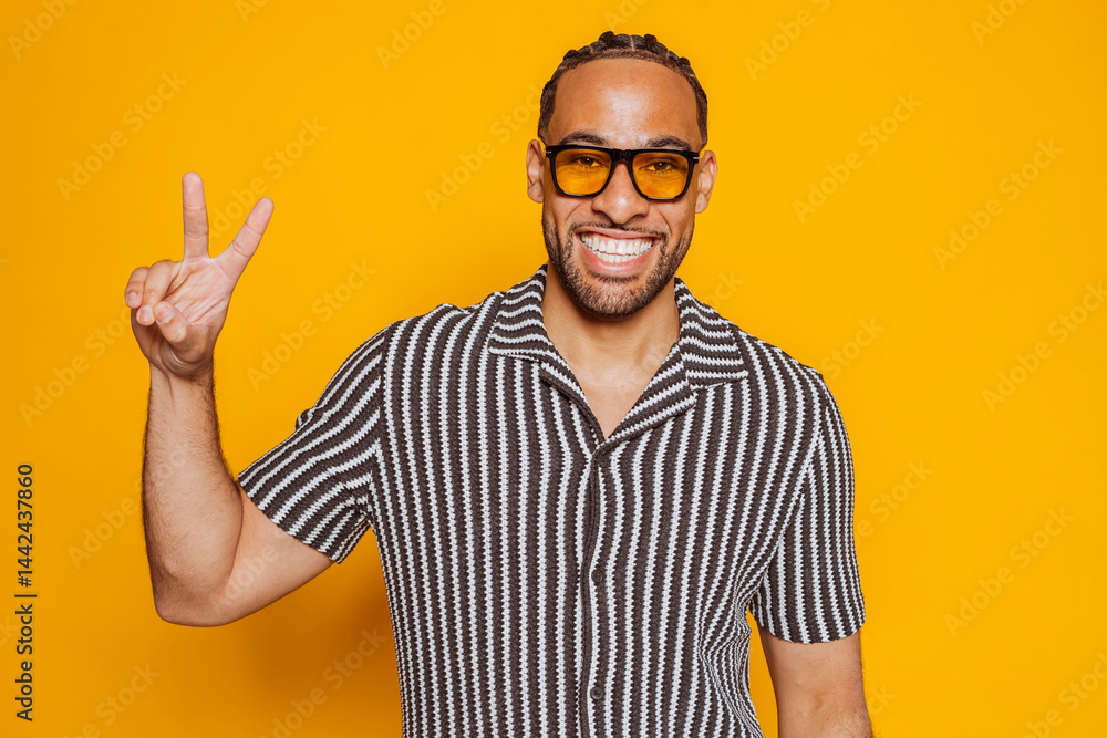 Naklejka premium Portrait of smiling young multiracial man wearing sunglasses showing victory sign while standing against yellow background at studio. 