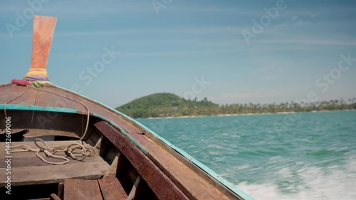 Bow of a wooden longtail boat cruising on beautiful, clear, turquoise water under a bright blue sky, with a green hilly island in the distance, handheld shot.