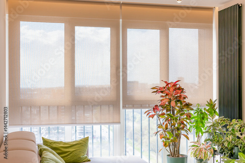 Automatic roller blinds cover large windows in a bright modern room, softly diffusing natural light. Beige transparent screen fabric for roller shades in smart home. Vertical radiator on the wall.