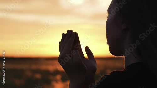 Woman praying with folded hands at sunset silhouette. Religious pious orthodox christian spiritual believing faithful girl at morning dawn sunrise reading prayer bible turning to god jesus christ.