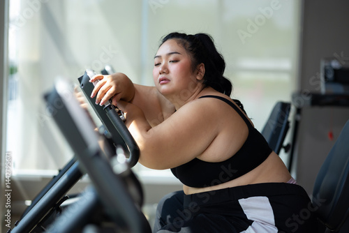 Tired fat asian woman on cycling. beautiful young chubby overweight woman wearing sporty fitness clothes doing exercise indoors at gym fitness sport club, body and health care
