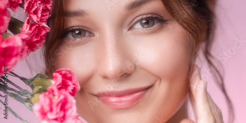 Obraz na płótnie Close-up of a smiling young Caucasian woman with pink carnations on soft pastel background. Ideal for beauty, skincare, floral fragrance, wellness, and feminine lifestyle promotions. w salonie