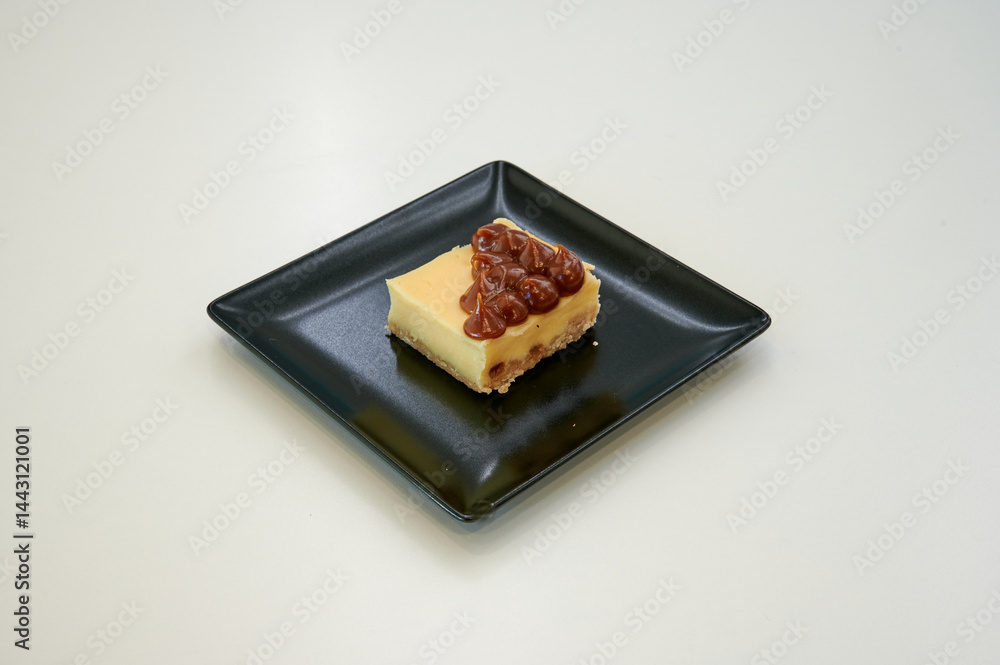 A slice of cheesecake with nuts on top. It is on a black plate. The plate is on a white table