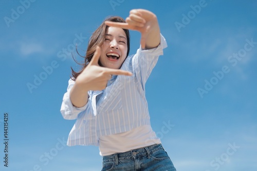 A cheerful woman wearing a casual shirt and jeans standing in front of the blue sky background with copy space.
