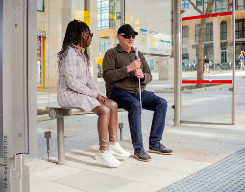 Blind man with a guide cane speaks with a woman at the bus stop.