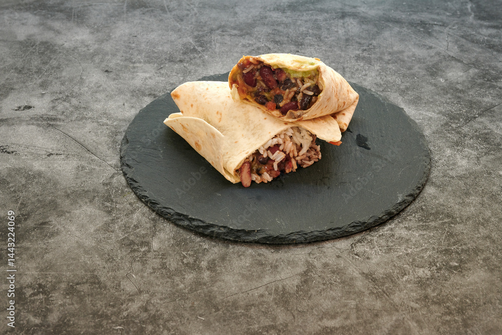 Two burritos on a black plate. One is open. The other is closed. The plate is on a gray surface