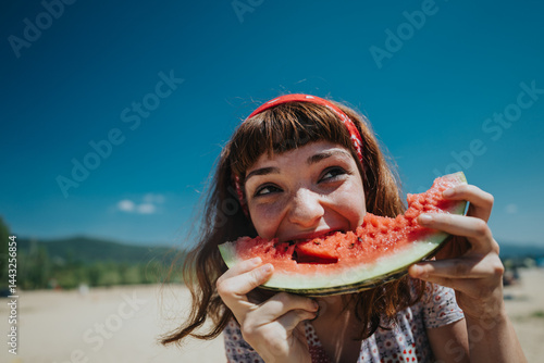 Obraz na płótnie A cheerful young woman bites into a watermelon slice on a bright summer day, capturing the joy of nature and outdoor fun near a scenic beach and lake environment. w salonie