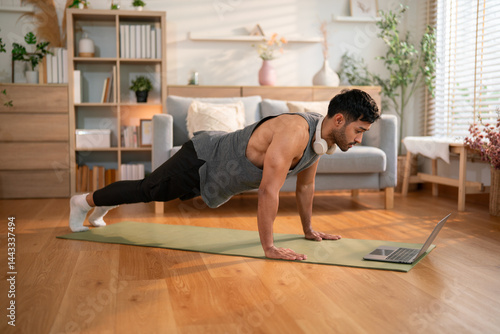 Muscular man wearing headphones in high plank pose on mat, engaging in strength training while watching an online video at home.