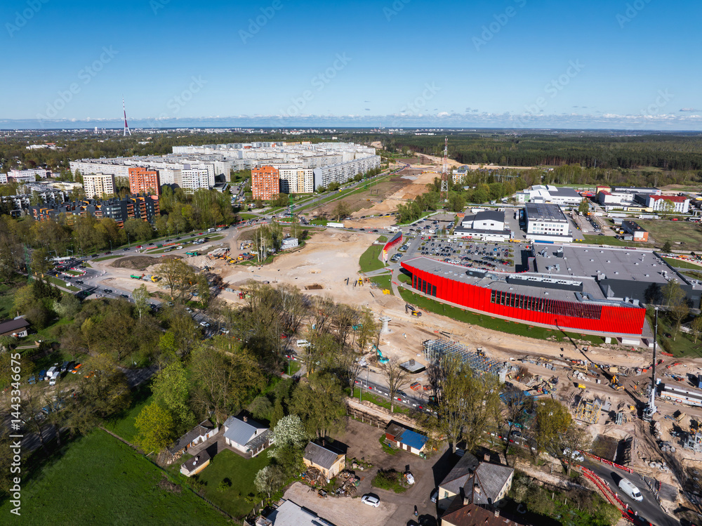 Aerial view of an active construction site in Riga, Latvia, with machinery, a red building, residential blocks, green spaces, and the Riga TV Tower visible.