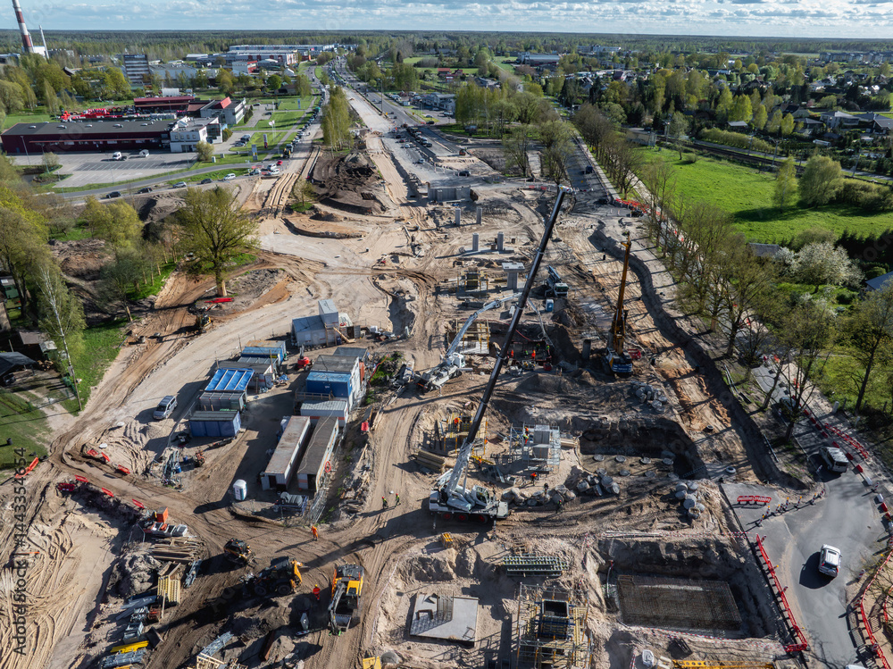 Expansive construction site in Riga with cranes, excavators, and temporary facilities, surrounded by urban and green spaces under a clear sky.