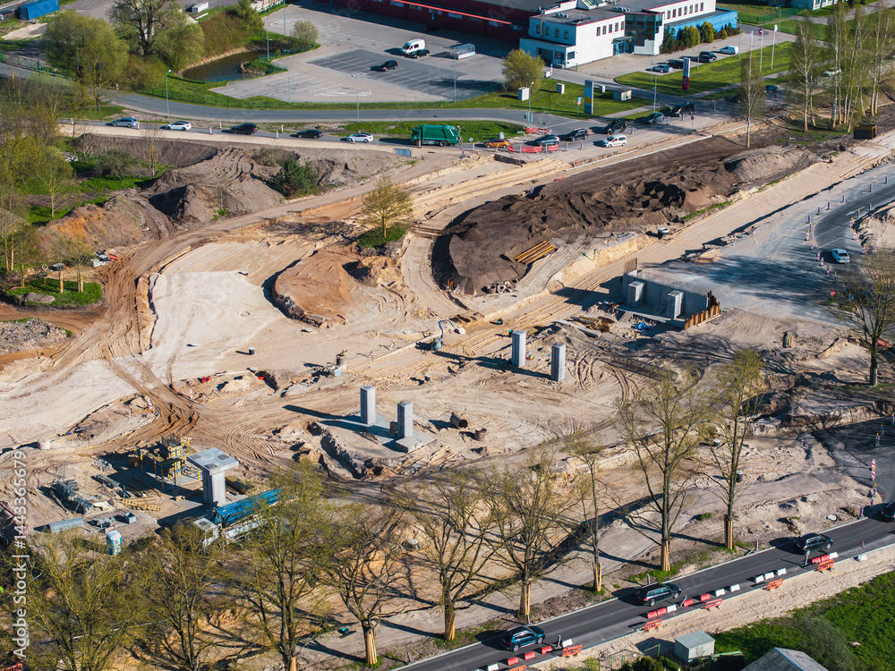 Aerial view of a construction site in Riga, Latvia, showing concrete pillars, roads with traffic, green spaces, and urban infrastructure in the background.