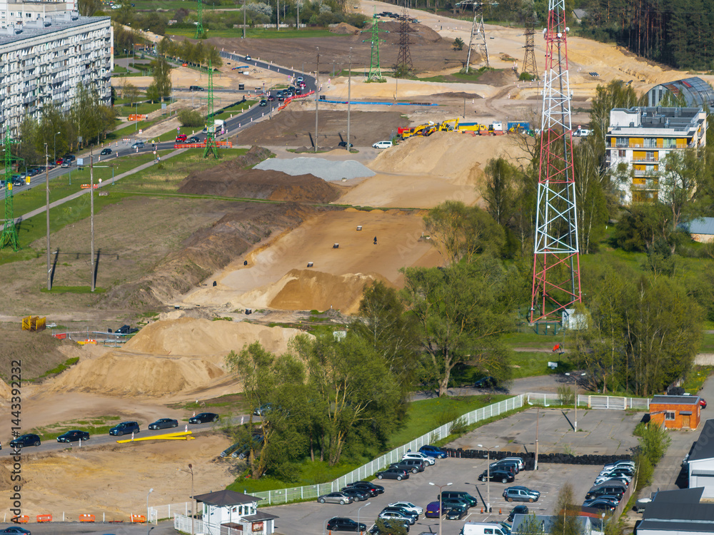 Active construction site with sand mounds, vehicles, workers, a red and white tower, residential buildings, parking lot, road traffic, and distant forest.
