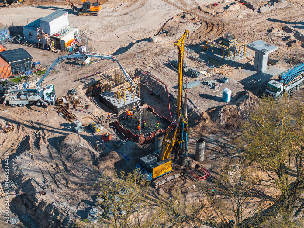 Active construction site with a yellow drilling rig, workers, machinery, and materials. Trees and scattered equipment frame the infrastructure project.