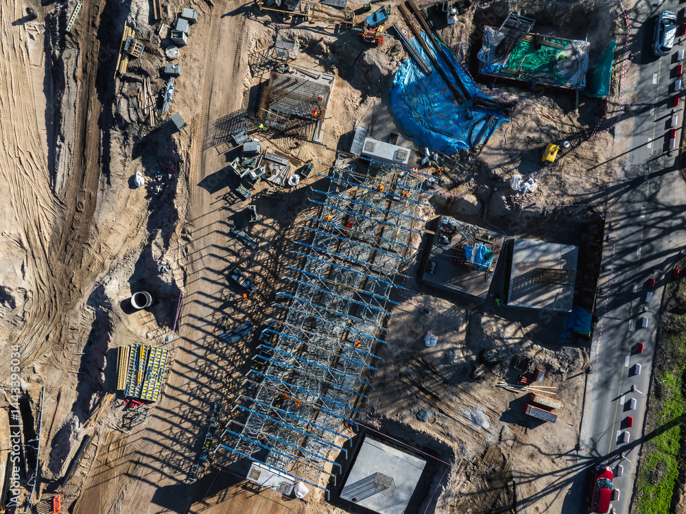 Aerial view of a construction site in Riga, Latvia, featuring a steel framework, workers, machinery, dirt roads, and visible progress on infrastructure.