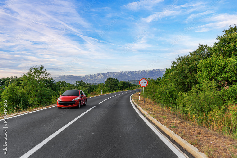 A red car drives along a winding mountain road surrounded by lush greenery, under a bright blue sky with a visible no overtaking traffic sign.