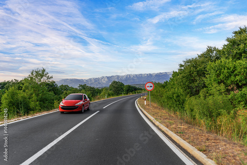 Obraz na płótnie A red car drives along a winding mountain road surrounded by lush greenery, under a bright blue sky with a visible no overtaking traffic sign. w salonie