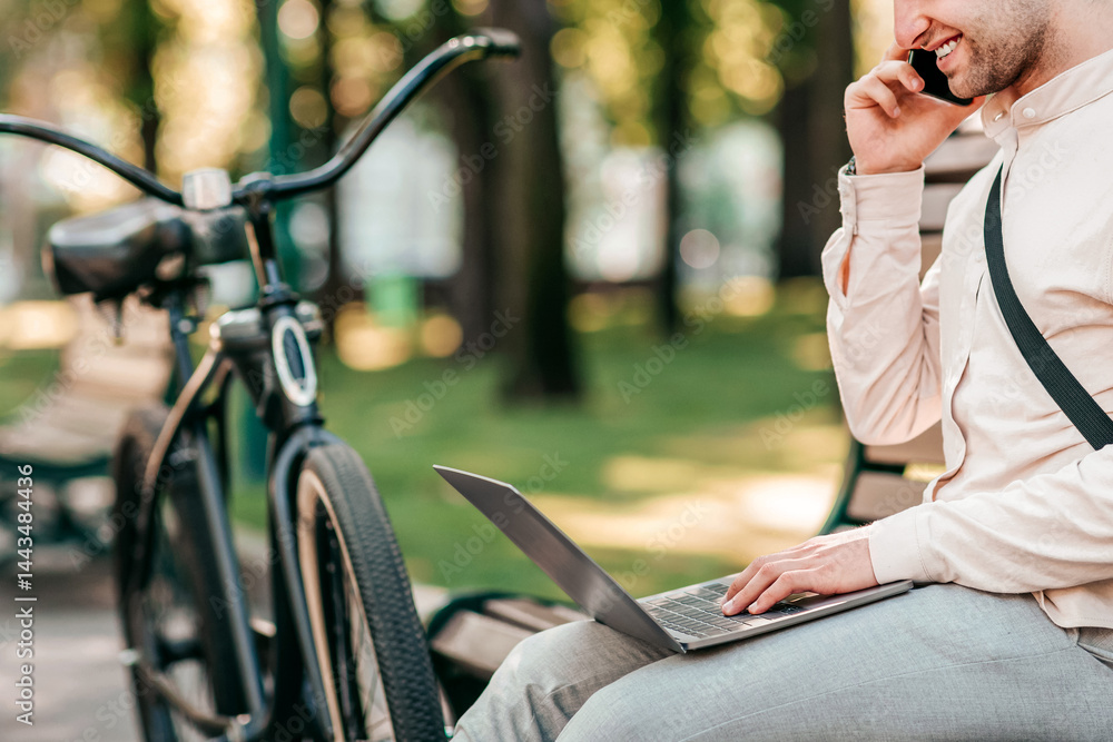 A young man seated on a bench in a park engages in a phone call while using a laptop. His bike rests nearby, showcasing a blend of technology and lifestyle in a relaxing outdoor setting. A young man seated on a bench in a park engages in a phone call while using a laptop. His bike rests nearby, showcasing a blend of technology and lifestyle in a relaxing outdoor setting.