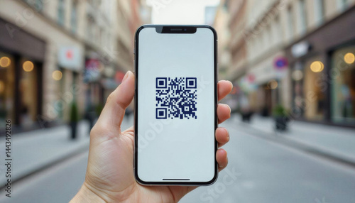 A hand holds a smartphone displaying a QR code against a blurred urban streetscape with shops and buildings.