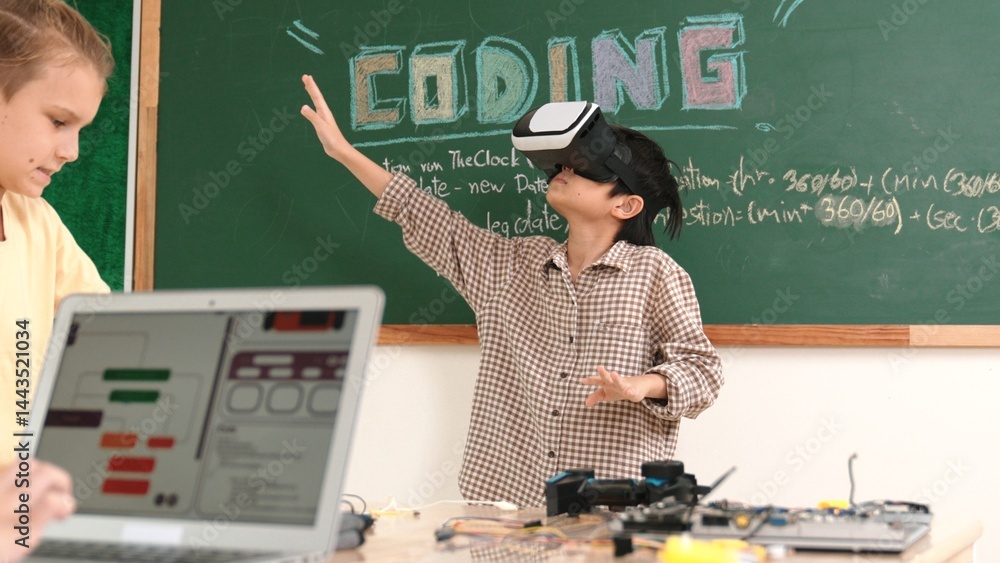Asian boy wearing VR headset to enter metaworld at table with board while girl coding system. Diverse student working together to develop program and writing engineering code. Pedagogy.