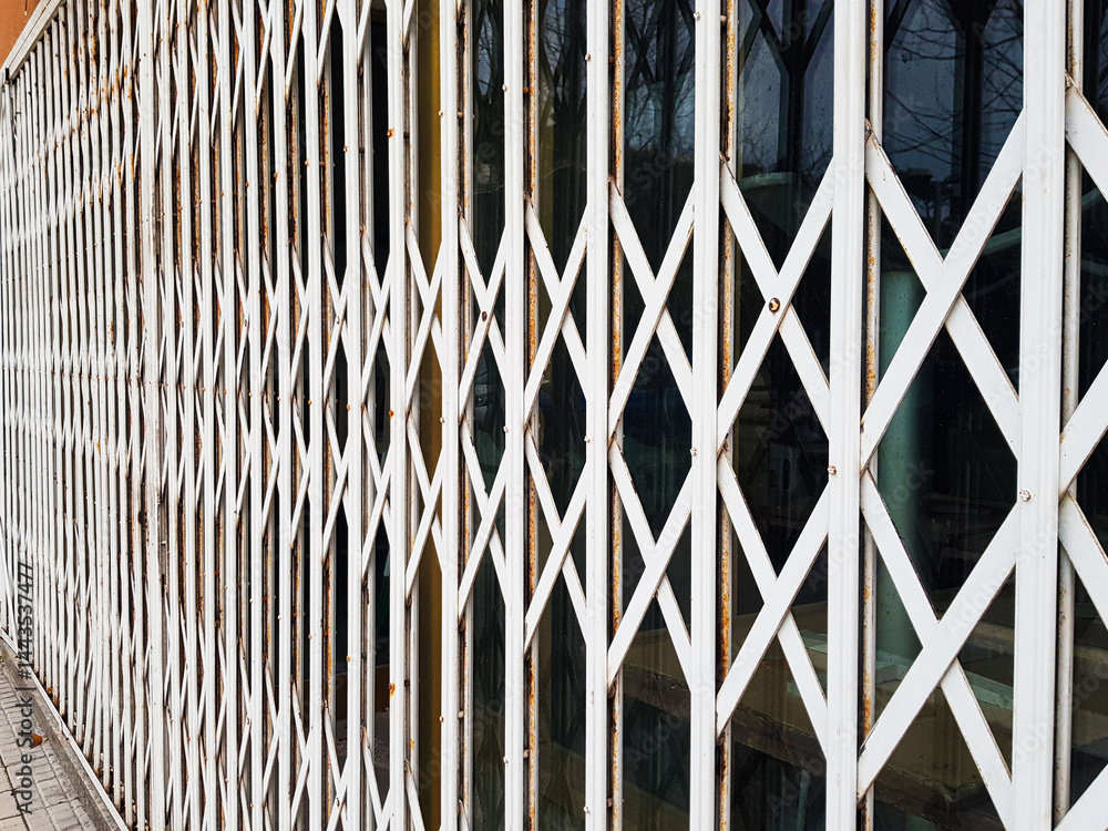 An intricate design of white metal grating covers the storefront of a business, reflecting the light of day and highlighting security features while indicating closure