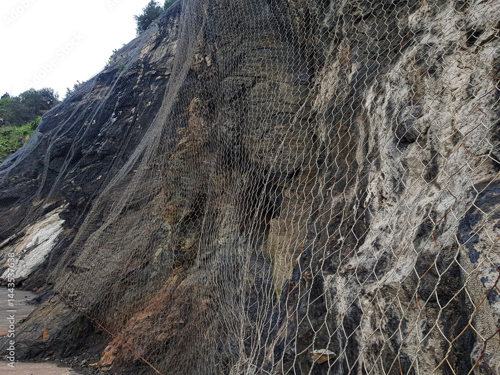 A coastal road features a steep rock face secured with netting, designed to protect against landslides. The area is rocky and appears to be near a body of water