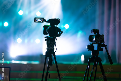 Two video cameras on tripods are positioned on a stage with dramatic lighting and colored spotlights, creating a cinematic and production-ready atmosphere.