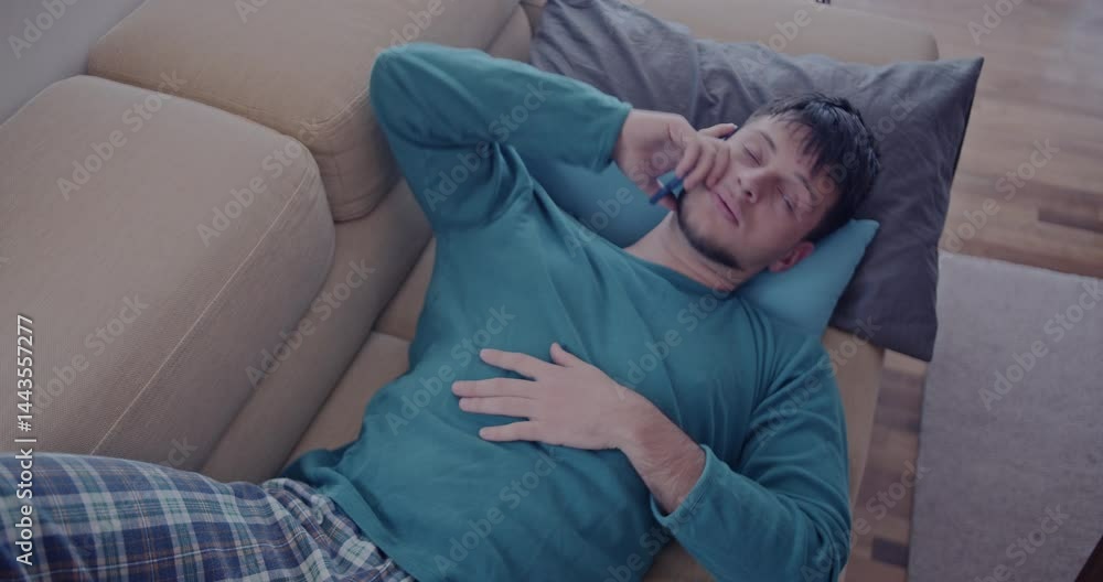 A young man relaxes on his couch, wearing pajamas and talking on his cell phone. He appears calm and comfortable. A young man relaxes on his couch, wearing pajamas and talking on his cell phone. He appears calm and comfortable.