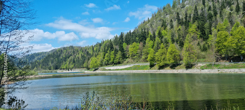 Lake Bajer in Gorski Kotar is captured on a sunny day with reflections of the surrounding lush green, forested hills and a partly cloudy blue sky on its calm surface