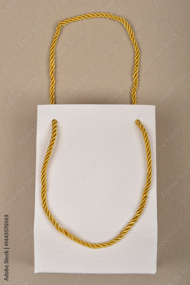 A small, white paper gift bag with shiny gold twisted rope handles is presented against a plain beige background