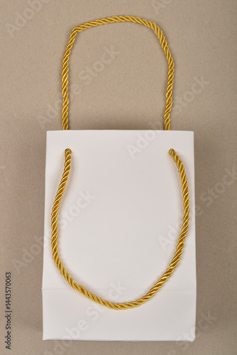Obraz na płótnie A small, white paper gift bag with shiny gold twisted rope handles is presented against a plain beige background w salonie