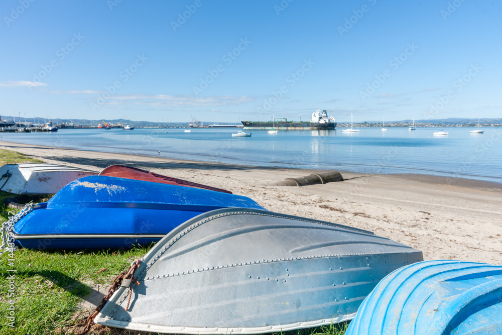 Group of upturned dinghies pulled up on grass edge to beach with boats and port activity in background