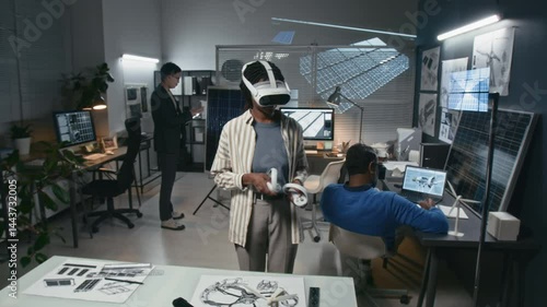 Push in shot of female innovator using virtual reality headset and controllers, developing renewable solutions in modern office, colleagues working on solar panels and wind turbines