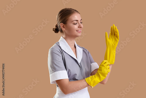 Young housemaid putting rubber gloves on beige background