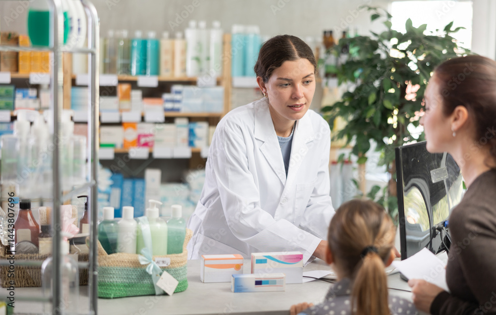 Obraz premium Professional young pharmacist using computer to check medication stock, politely explaining treatment options, and processing order for female customer with tween daughter