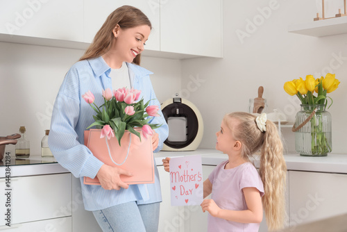 Cute little girl greeting her mom for Mother's Day with tulips and postcard in kitchen