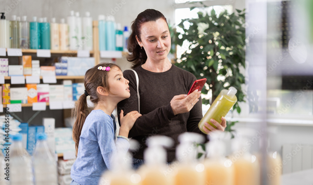 Woman stands with daughter in a pharmacy and makes purchases using an online application. Customer with a child scans the barcode of a plastic shampoo bottle to read the instructions on the website