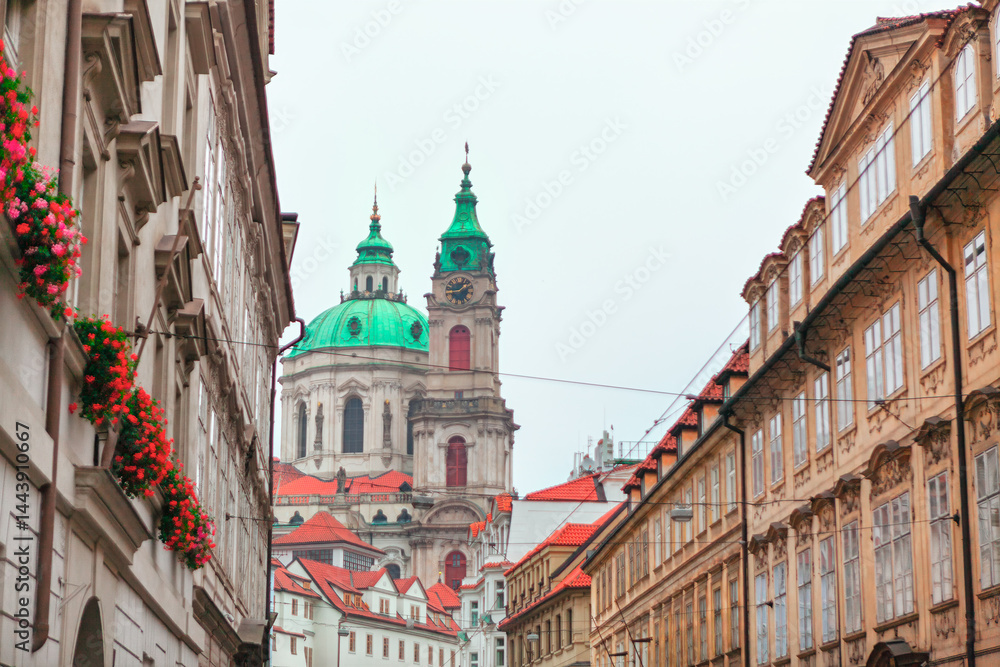 Church of Saint Nicholas, Baroque church in Prague Lesser Town, towers over a narrow street with its green domes, framed by historic buildings. Red flowers in window boxes add vibrant touch to scene