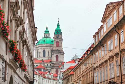 Church of Saint Nicholas, Baroque church in Prague Lesser Town, towers over a narrow street with its green domes, framed by historic buildings. Red flowers in window boxes add vibrant touch to scene