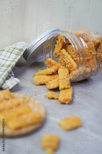 an open jar filled with cheese cookies that was lying down and the cookies came out partially, with blurred cheese cookies on wooden coaster and gingham fabric