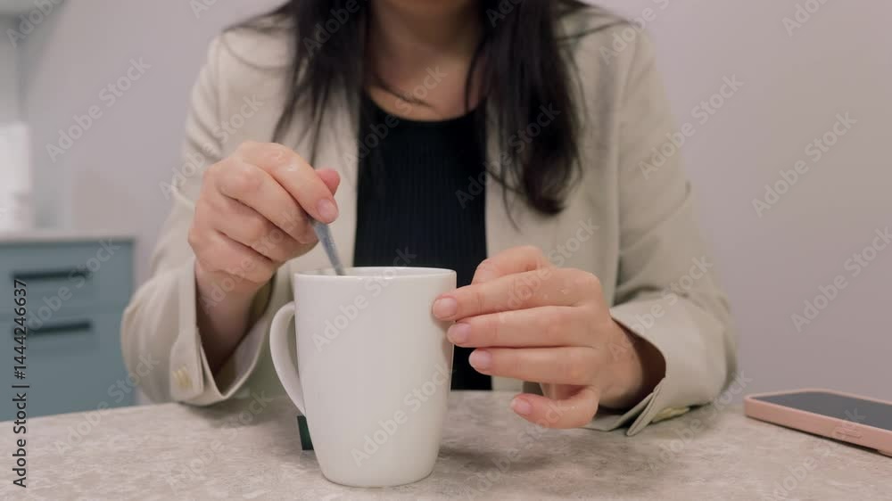Close up of female hands stirring coffee in white mug on office desk, with smartphone lying nearby, suggesting a moment of relaxation during workday Close up of female hands stirring coffee in white mug on office desk, with smartphone lying nearby, suggesting a moment of relaxation during workday