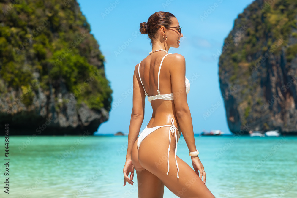 Confident young woman with glowing skin and perfect figure standing in white bikini, back view, admiring turquoise water at Maya Bay, Thailand, concept of luxury travel, fashion, and high quality